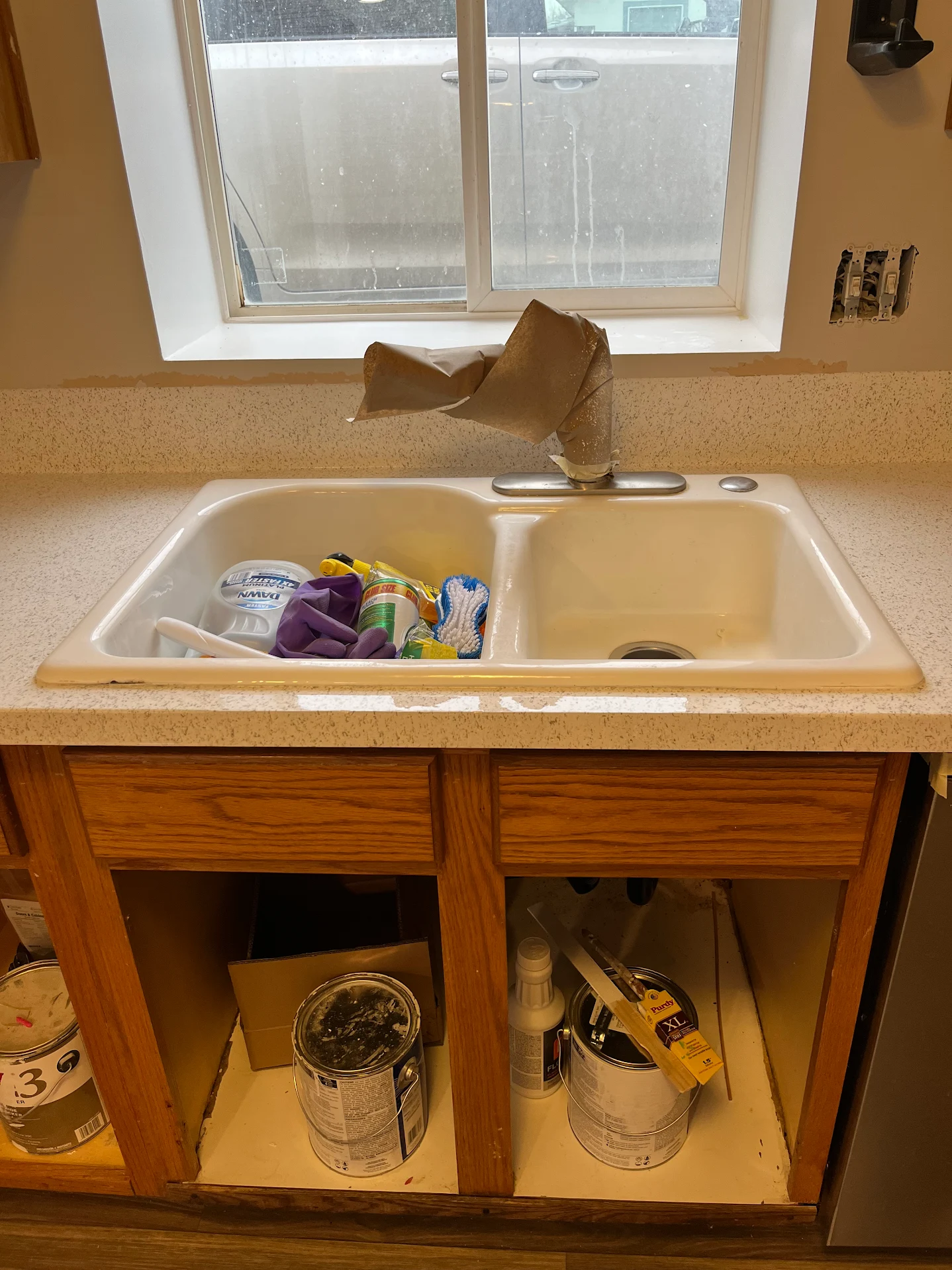 Kitchen sink with a covered faucet and cleaning supplies in the left basin, beneath the sink are open cabinets storing paint cans and cleaning products.
